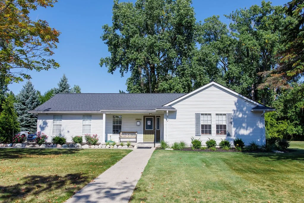 A white single-story house with a dark gray roof, surrounded by green trees and a well-maintained lawn, with a sidewalk leading to the front door.