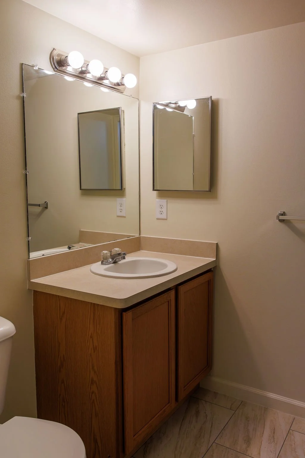 Bathroom vanity with single sink, beige countertop, wooden cabinet, two mirrors, light fixture with four bulbs, and a beige tiled floor.