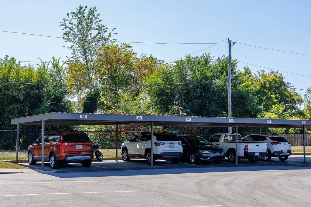 A parking area with parked cars, including an orange SUV, a white SUV, a black sedan, a white pickup truck, and another car, under a long metal canopy with labeled sections, against a background of trees and power lines.