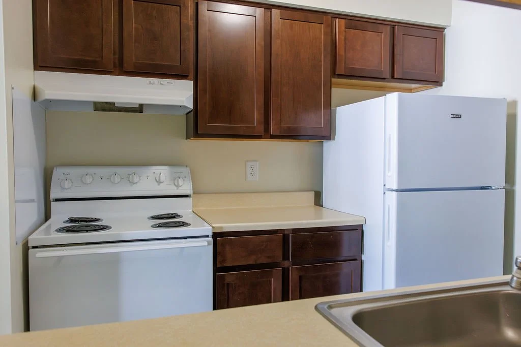 Kitchen with brown upper cabinets, white refrigerator, white stove, beige countertop, and stainless steel sink.