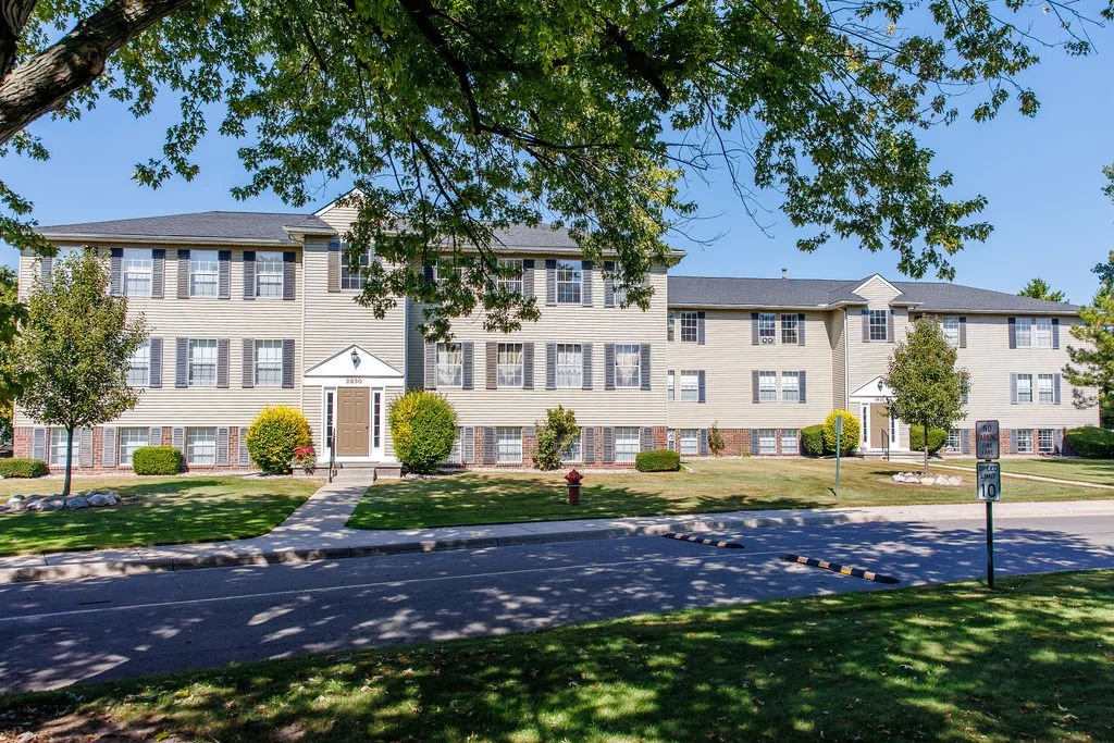 A multi-story residential apartment building with a well-maintained lawn and trees in front, under a clear blue sky.