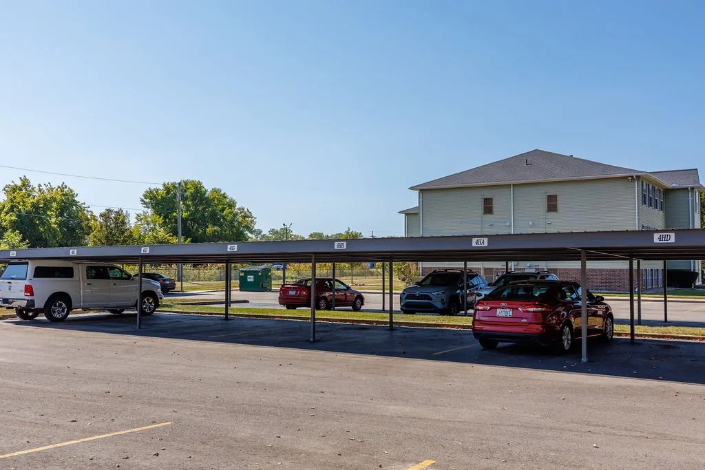Carport with vehicles parked underneath, situated on an asphalt lot with a residential building in the background.