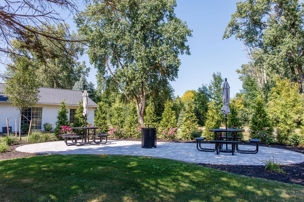 Outdoor patio area with round picnic tables, umbrellas, and a trash can, surrounded by trees and bushes, with apartments visible in the background.