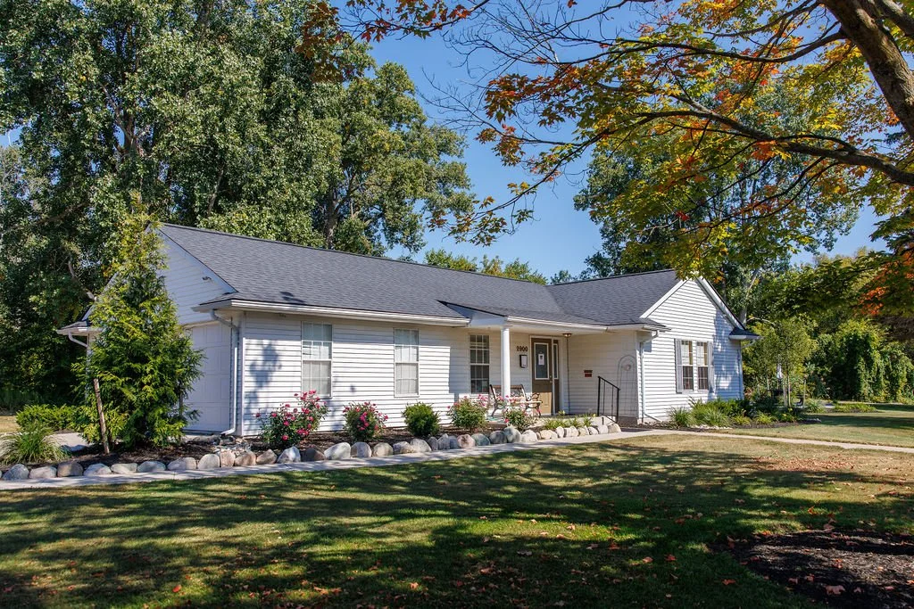 White house with a black roof, surrounded by trees and a green lawn, with a flower bed and stone border in front.