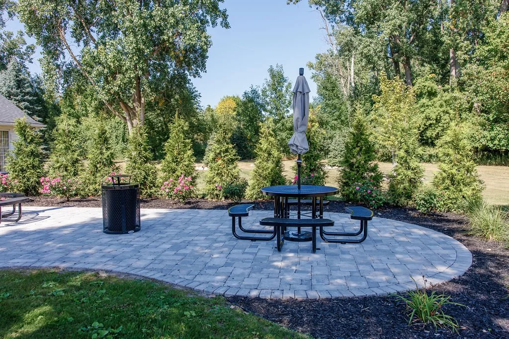 A backyard patio with a round picnic table with benches, an umbrella, a trash can, and floral bushes in the background.
