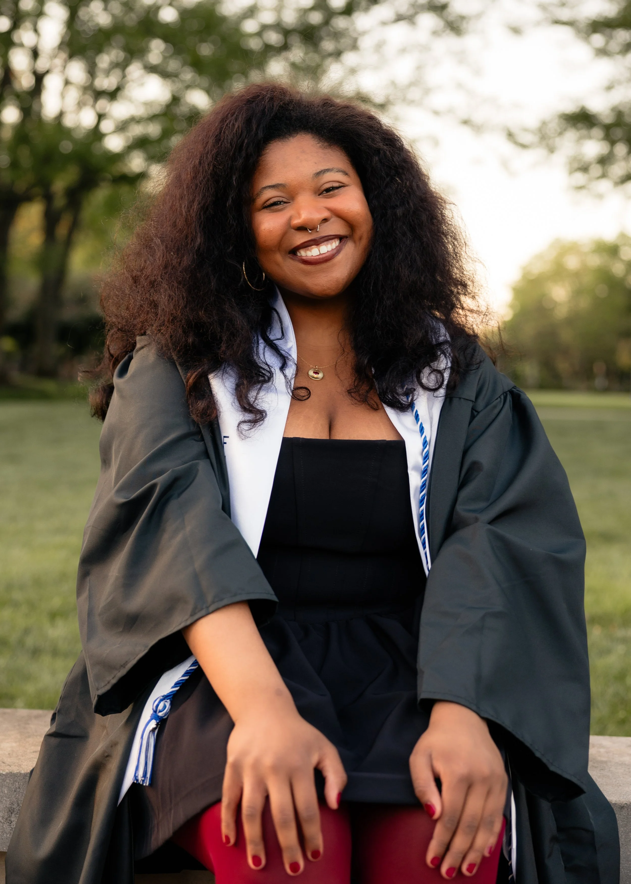 A smiling woman with curly hair and a septum piercing sitting outdoors, wearing a black dress and a graduation gown.