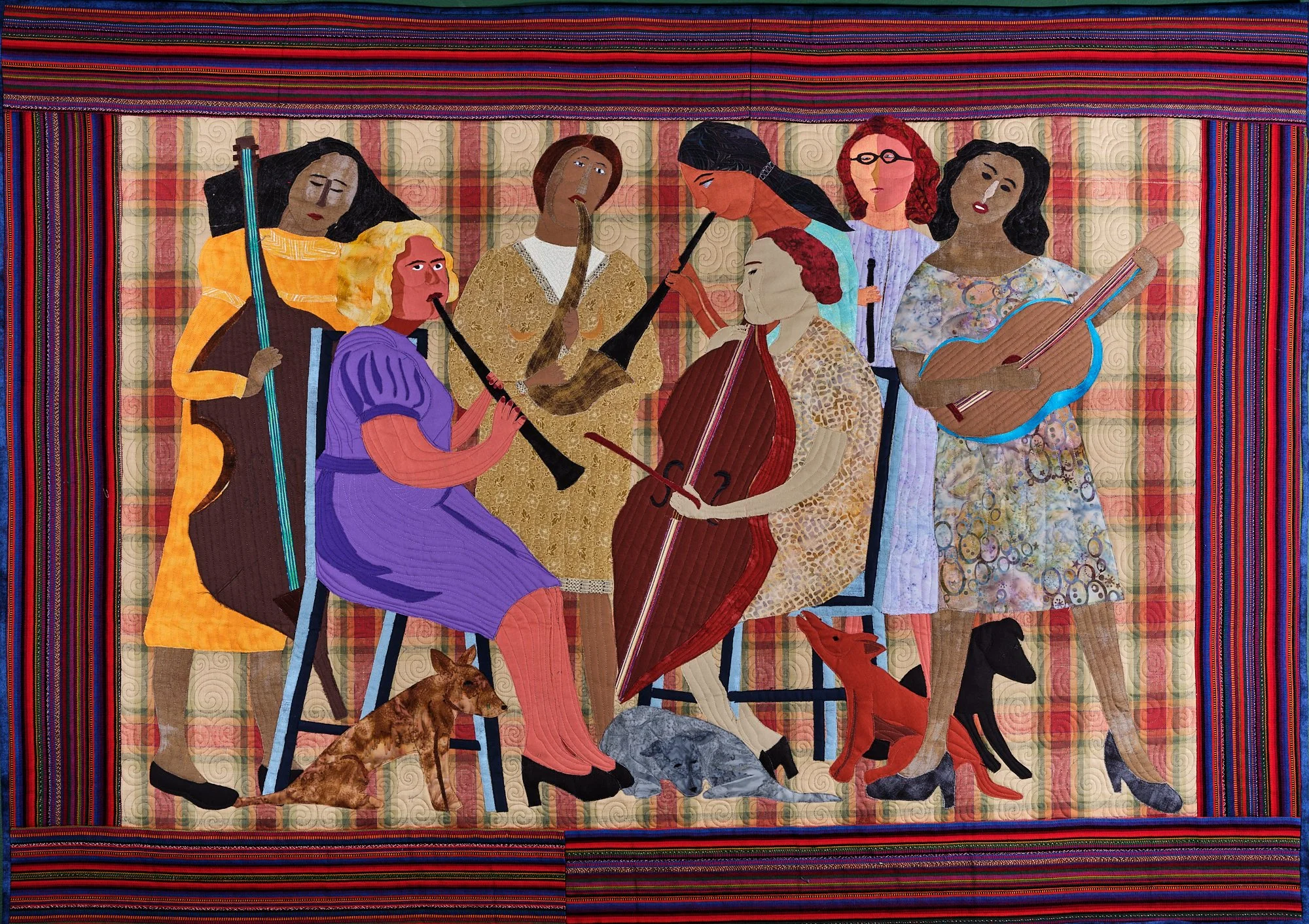 A colorful quilted artwork by Susan Zepeda depicts six women playing musical instruments, including a cello, guitar, recorders, and violin. Striped and plaid fabrics surround them as two cats sit contentedly on the floor.