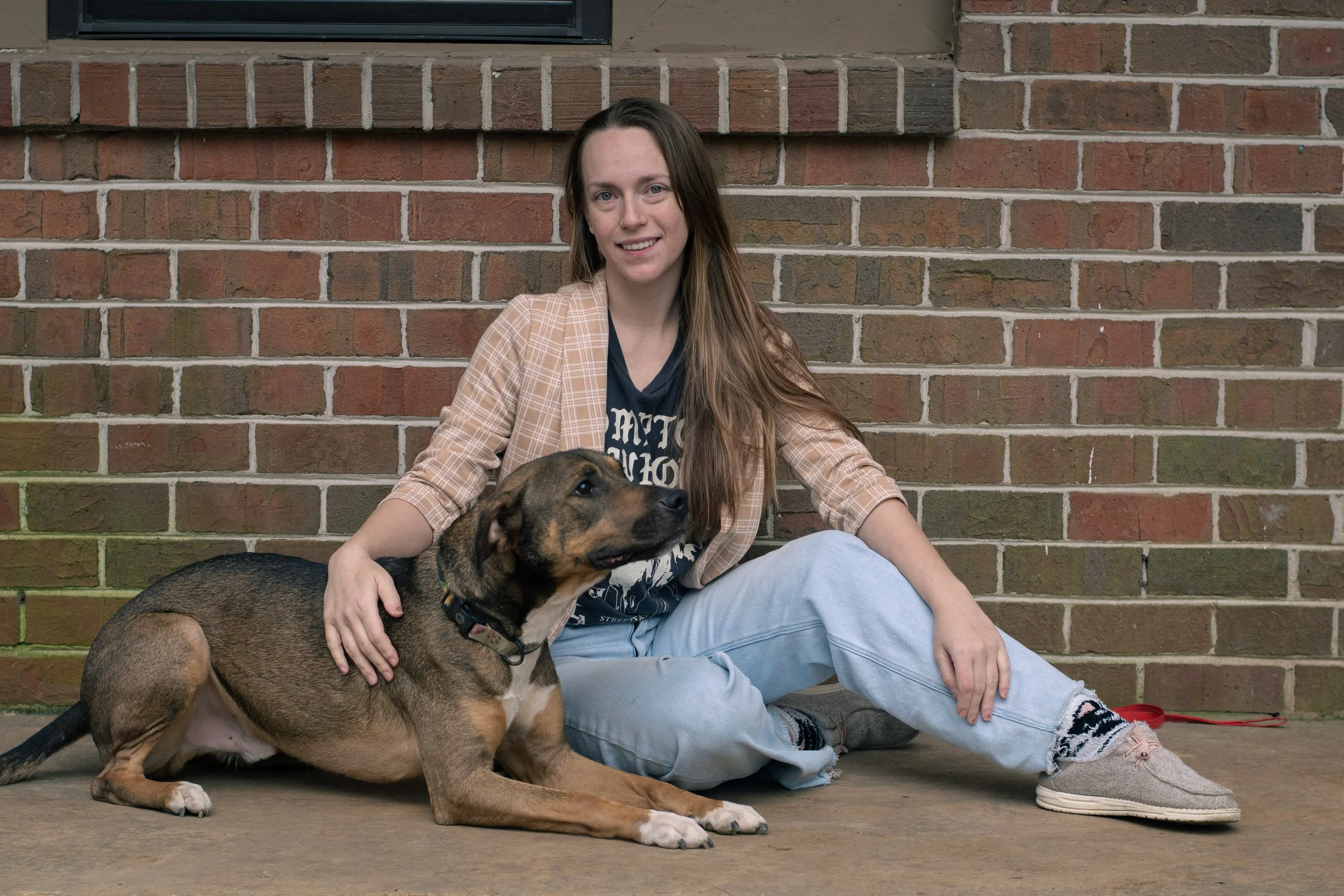 A young woman with long brown hair sitting on the ground against a brick wall, embracing a large brown and black dog lying beside her.