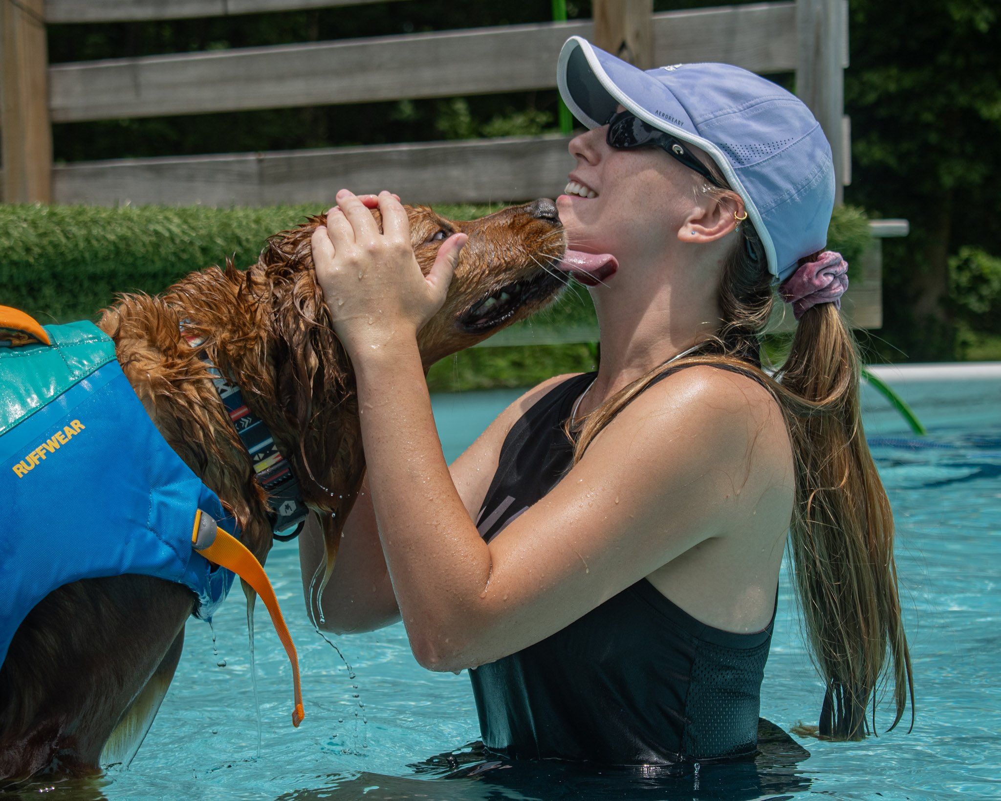 A young woman with long brown hair stands in a dock diving pool while a dog licks her face.