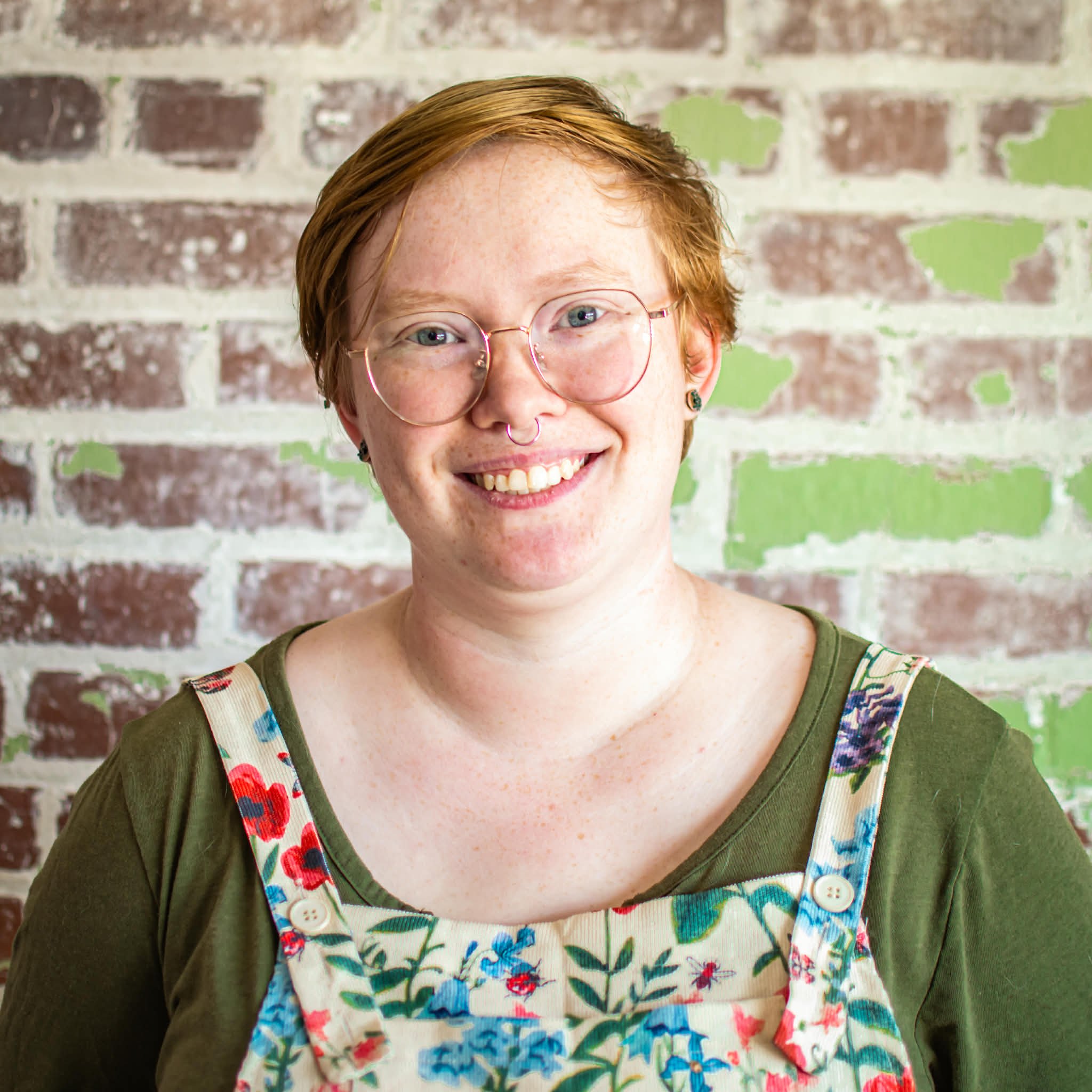 The head photographer for Red Pet Photography poses with short red hair, glasses, and a septum piercing, smiling in front of a brick wall painted with green and white patches, wearing a green shirt and a floral apron.