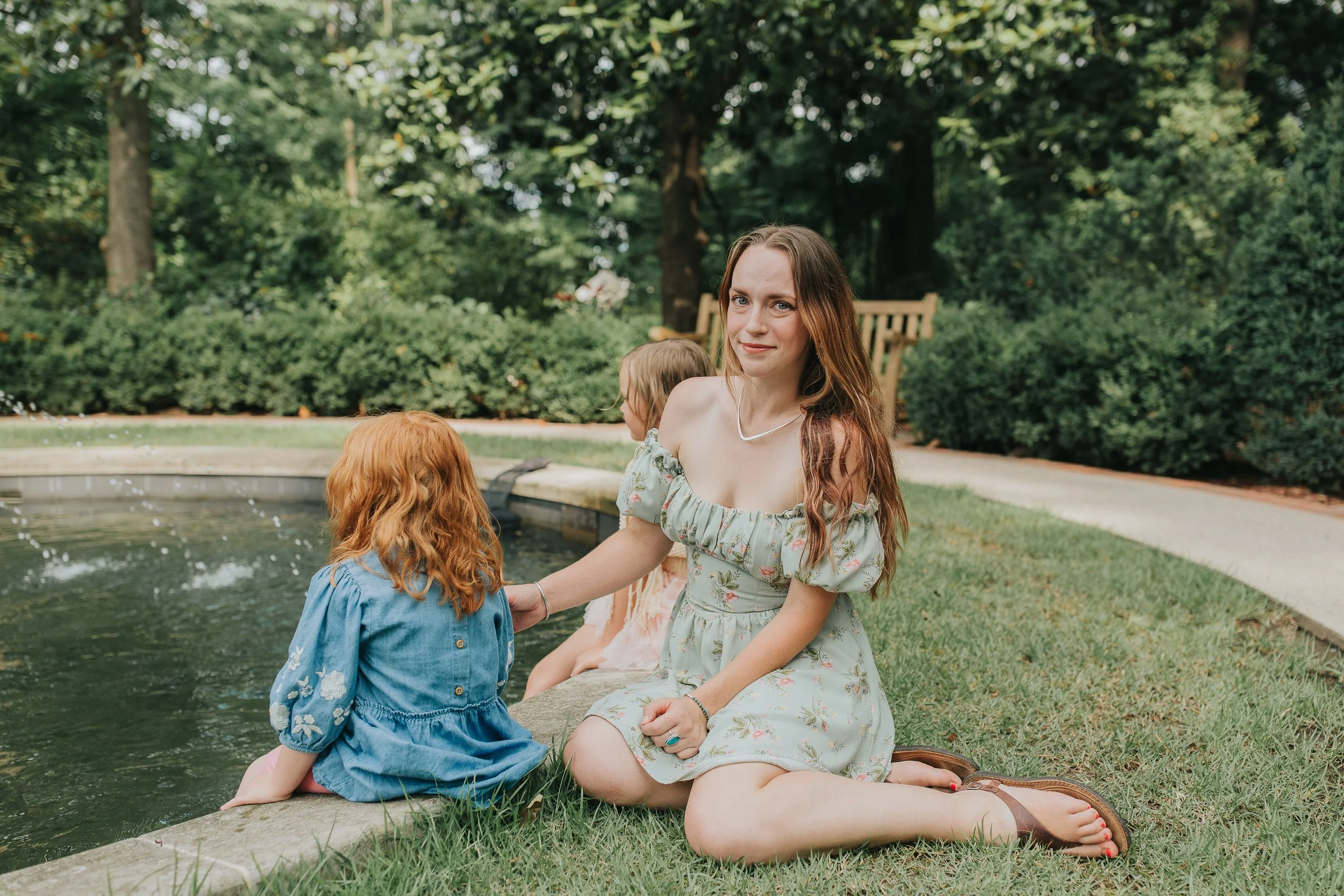 A woman with long brown hair, wearing an off-shoulder floral dress, sitting on the grass next to a small pond with two young children, with lush green trees and bushes in the background.
