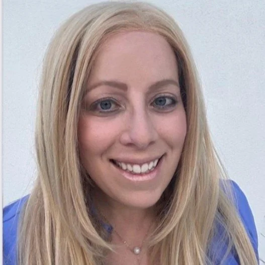 Close-up of a smiling woman with blonde hair, blue eyes, and a pearl necklace, wearing a blue blazer.