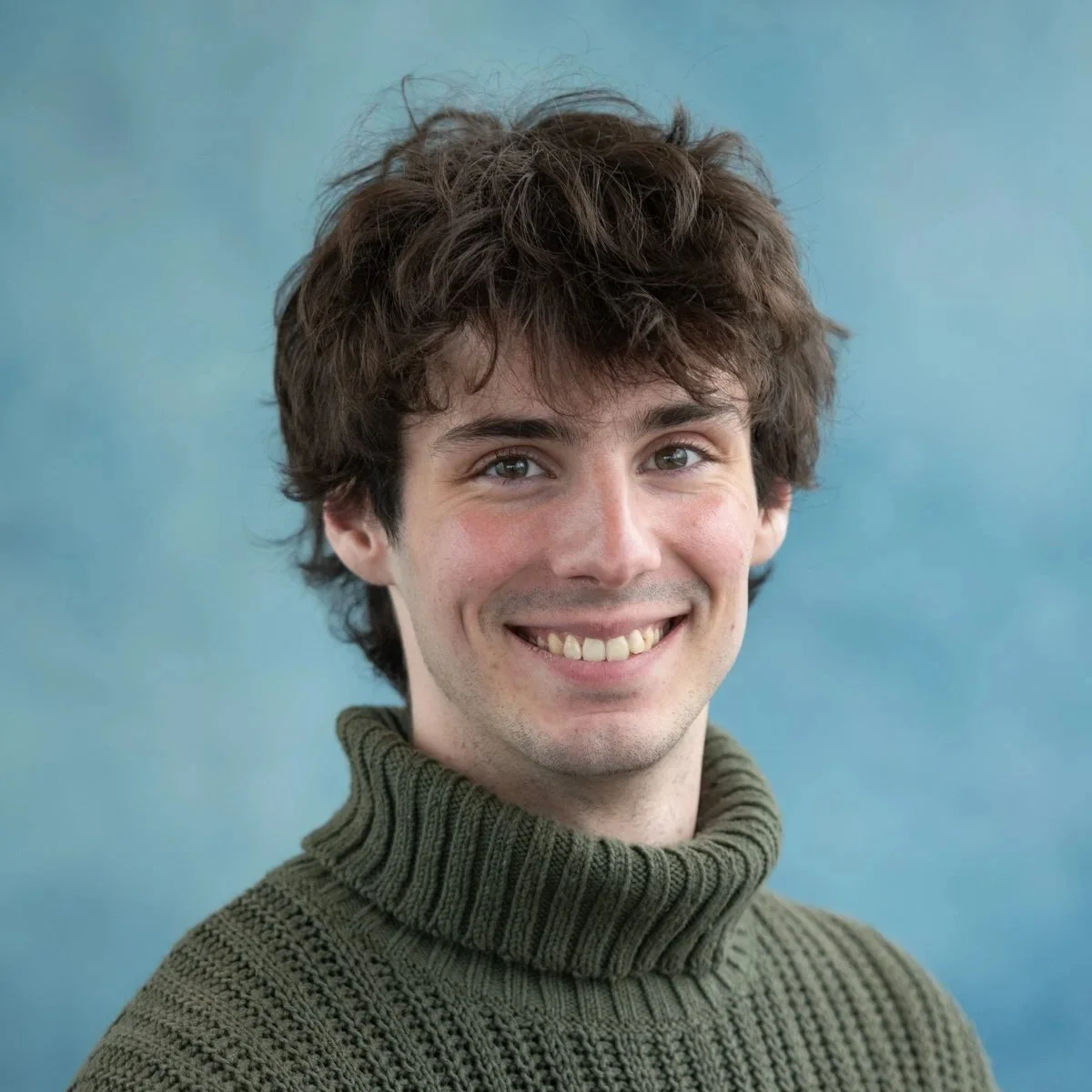 A young man with wavy brown hair and blue eyes smiling, wearing a green turtleneck sweater, standing against a blue background.
