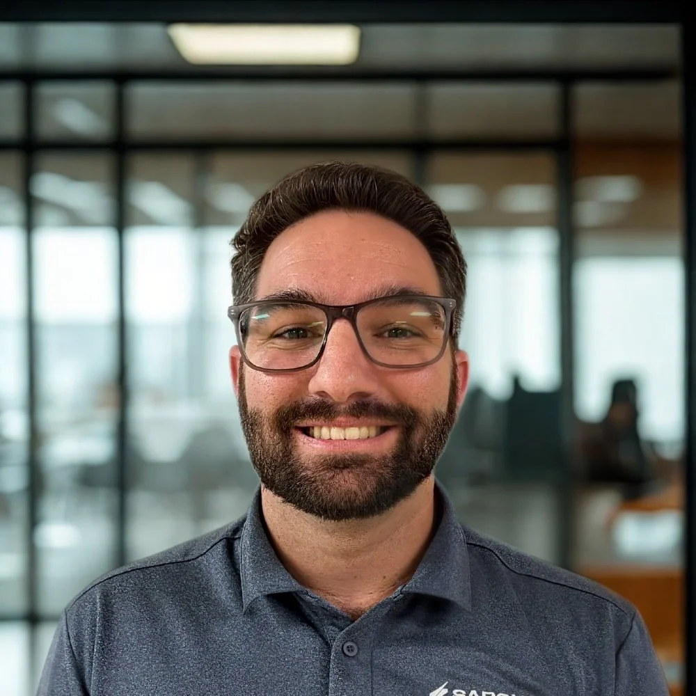 A smiling man with glasses and a beard, wearing a dark grey polo shirt, standing in an office setting with glass walls and blurred background.