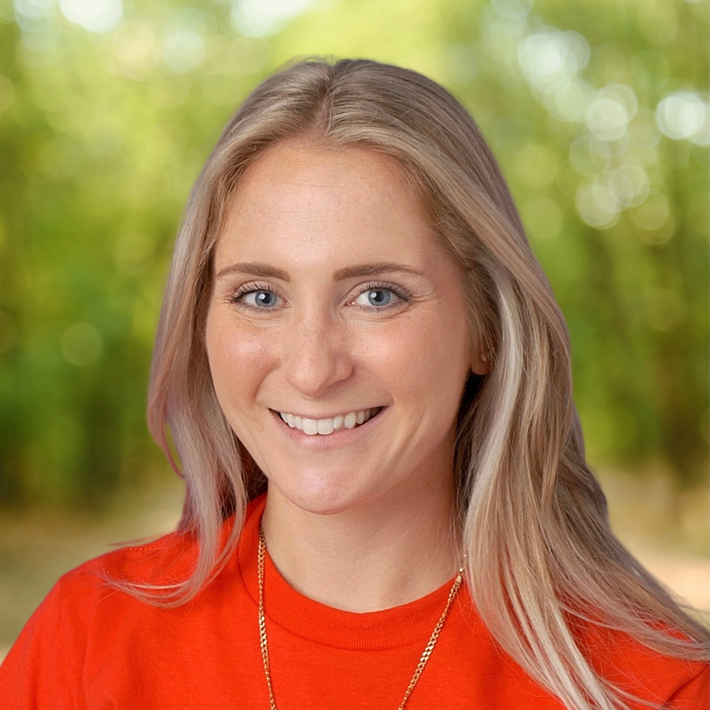 A young woman with blonde hair and blue eyes smiling, wearing a red shirt and a gold necklace, outdoors with blurred green trees in the background.