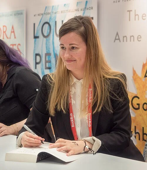 Picture of Tamsyn Muir. She is a woman with long blonde hair, wearing a black blazer and white blouse, is sitting at a table with a book and a pen, at a book signing event. She has a red lanyard around her neck with the words "Bookbub" on it.