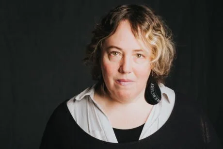 Portrait of a Kelly Link, who has short, curly hair, wearing a black top over a white shirt, against a dark background.