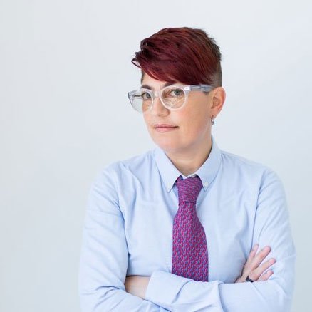 A man with curly dark hair, clear glasses, and a beard looks to the side with a slight smile. He is wearing a dark green sweater and a white shirt underneath, sitting in a bright room with a white wall and a plant in the background.