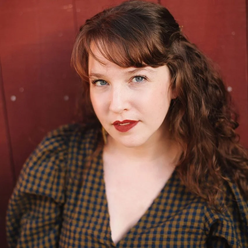 A woman with curly hair wearing glasses, earrings, and a striped shirt, smiling in an indoor setting.