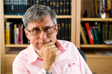 A man with gray hair and glasses resting his chin on his hand in front of a bookshelf.