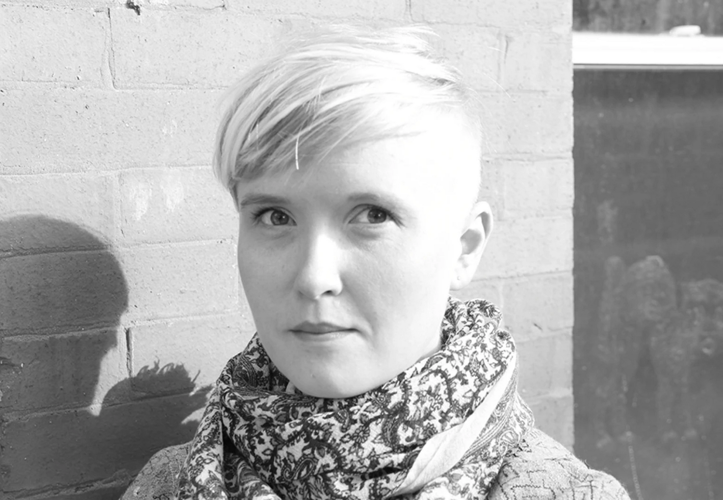 Photo of Jac Jemc, who has short light-colored hair, wearing a patterned scarf and coat, standing in front of a brick wall.