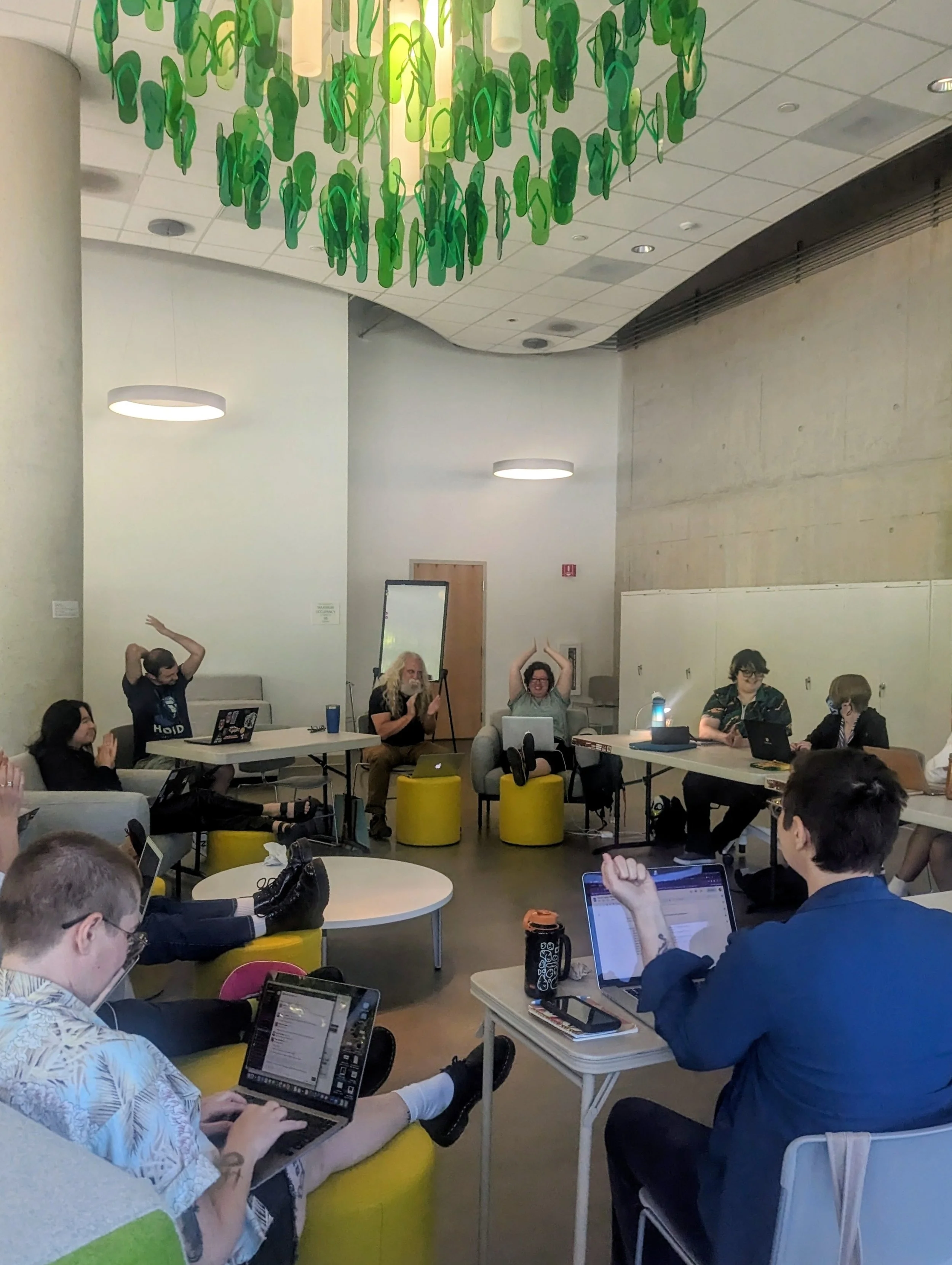 The 2025 Clarion class in a workshop in a modern indoor space, engaging in a group activity or meeting, with some individuals raising their hands or making gestures. A large green and yellow abstract art installation hangs from the ceiling.