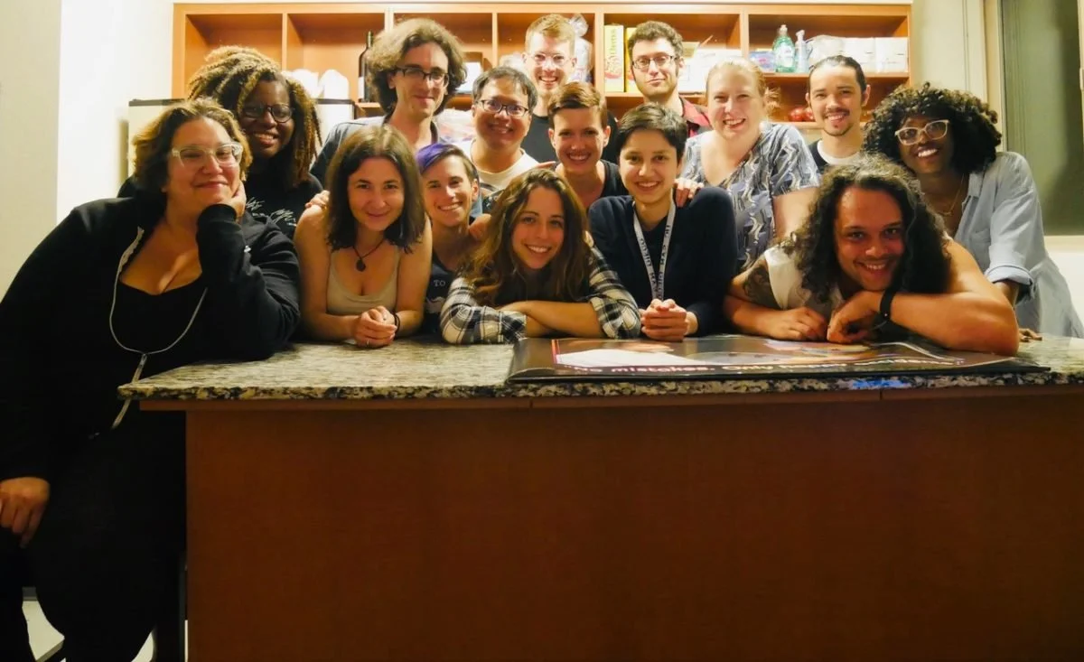 Group of sixteen diverse young adults in a kitchen, smiling and gathered around a granite counter, with cabinets and kitchen items in the background.