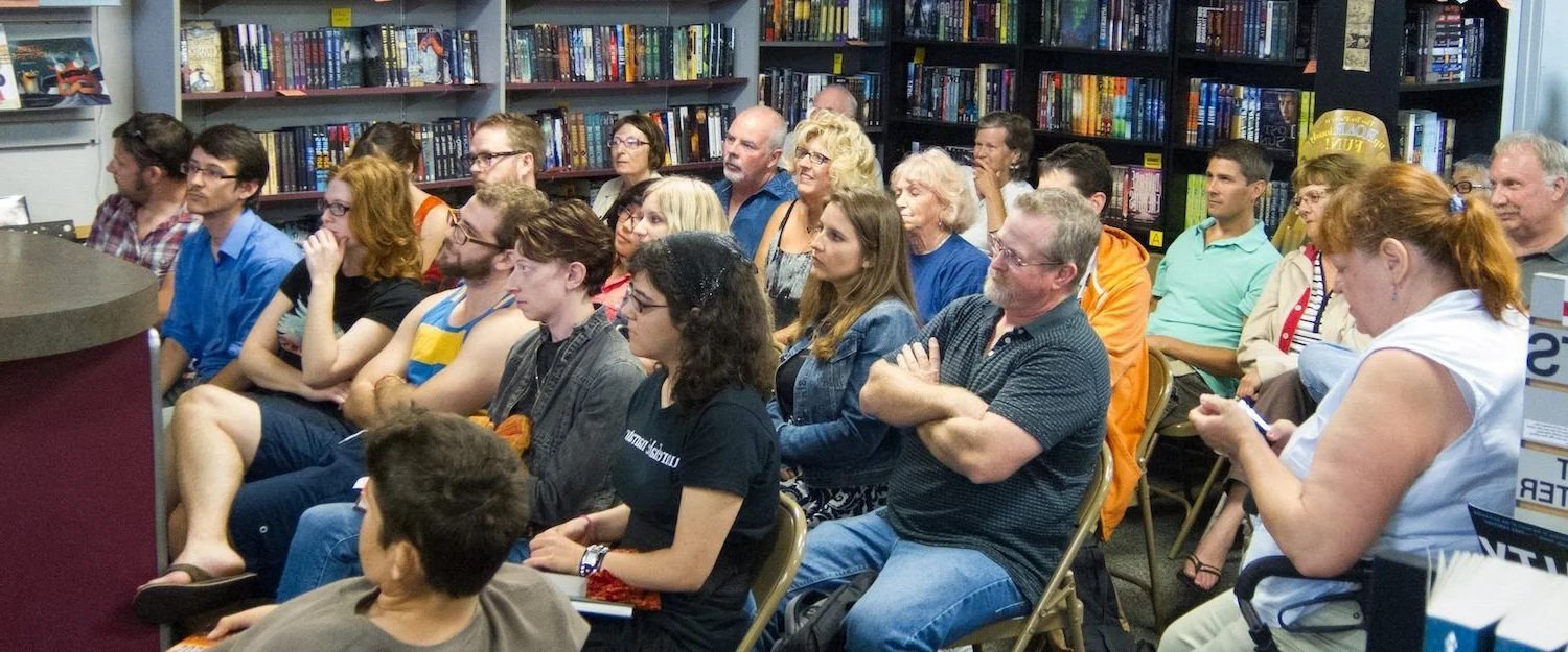 Audience seated in rows listening at a bookshop event with bookshelves in background.