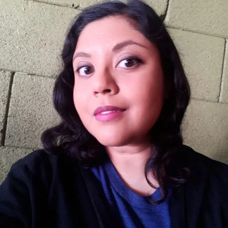 A man with dark curly hair, glasses, and a beard, wearing a dark green sweater, looking to the side with a slight smile, in an indoor setting with a plant in the background.