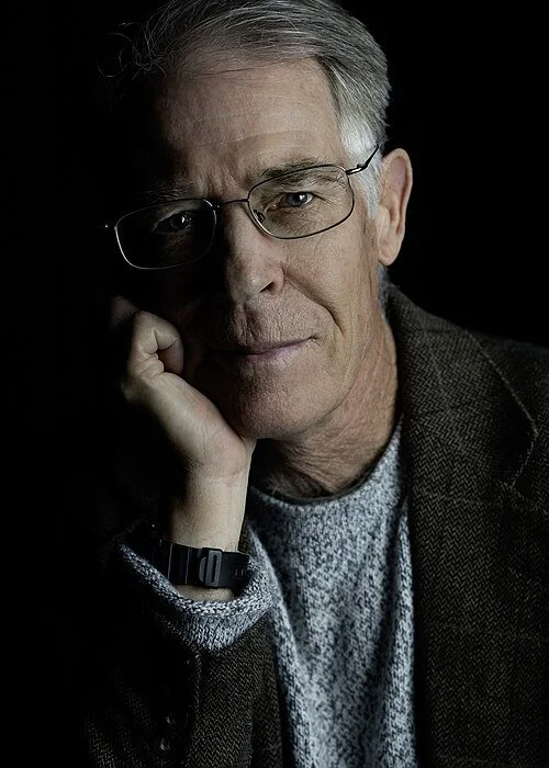 Close-up portrait of an older man wearing glasses, a gray sweater, and a brown jacket, resting his face on his hand with a thoughtful expression.