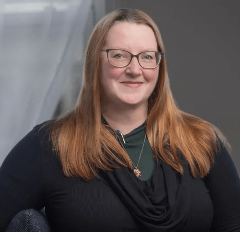 A woman with long red hair, glasses, and light skin, smiling at the camera. She is wearing a dark top, a necklace, and a green shirt underneath, sitting in a room with a gray background.