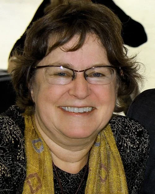 Smiling woman with short brown hair and glasses, wearing a patterned jacket and a yellow scarf.