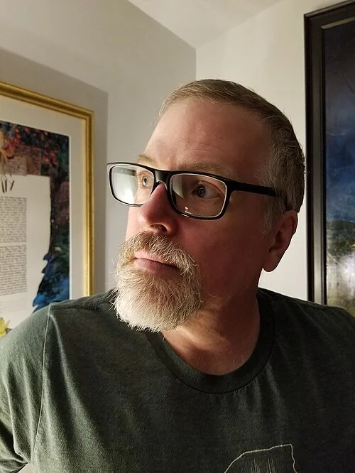 Jeff VanderMeer looking to the side, indoors with framed artwork in the background.
