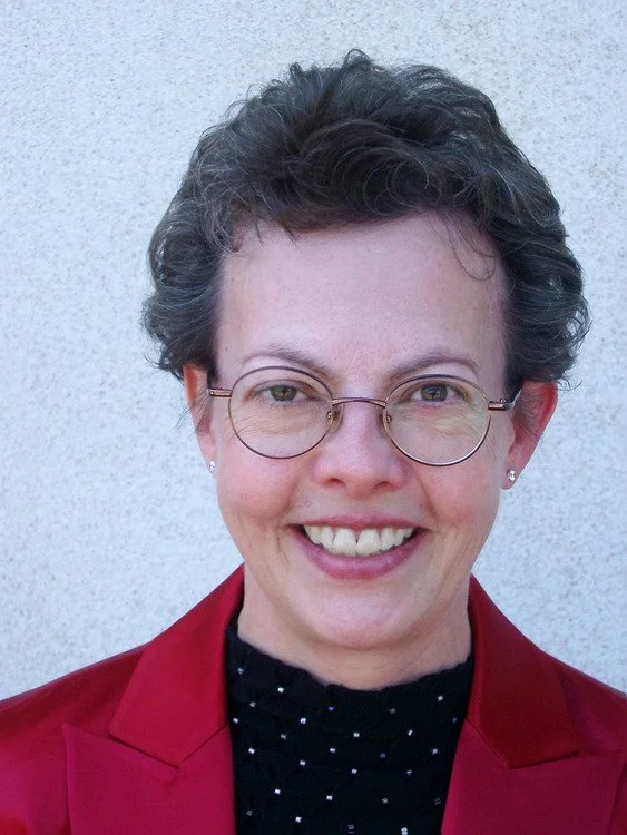 A woman with short curly gray hair, glasses, and small earrings, smiling, wearing a red blazer and black top with white polka dots, standing against a light-colored wall.