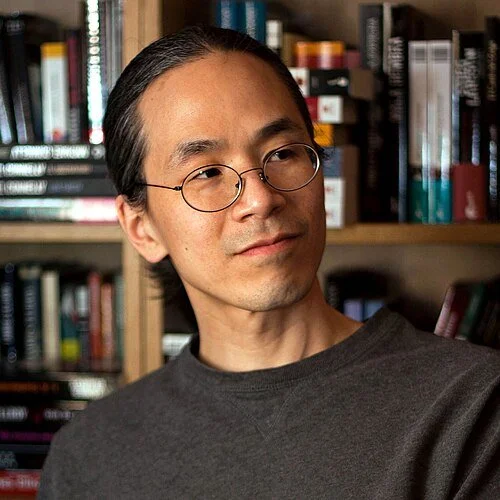 Picture of Ted Chiang. He is a man with glasses and long dark hair in a bun, wearing a gray shirt, standing in front of a bookshelf filled with books.