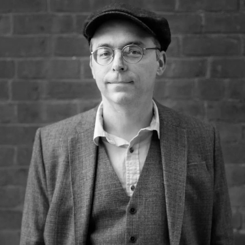 A smiling woman with black hair, wearing a blue top, in a lush green indoor garden or greenhouse.