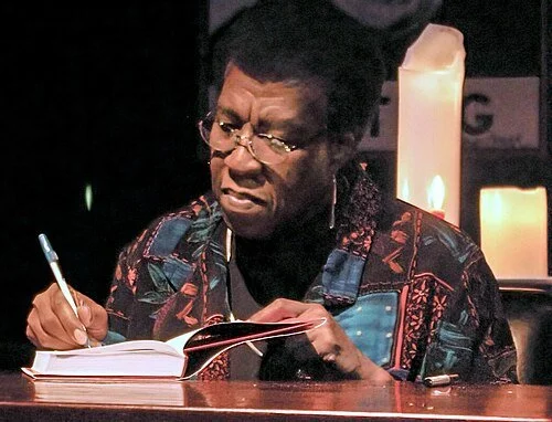 Octavia Butler signing one of her books at a table, with candles in the background.