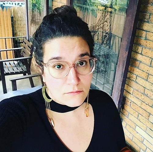 Picture of Carmen Maria Machado. She has dark hair in a bun, glasses, gold earrings, and a black choker necklace, taking a selfie outside on a porch with benches and a brick wall