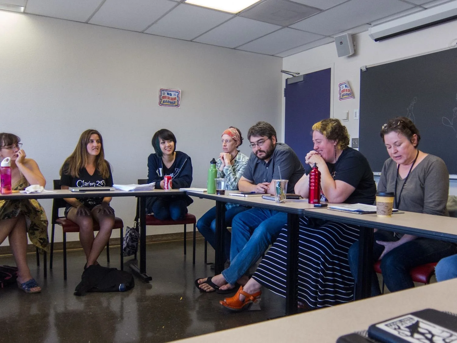 A group of seven people sitting at a conference table in a Clarion Workshop. There are notebooks, water bottles, and coffee cups on the table. A blackboard is visible in the background.