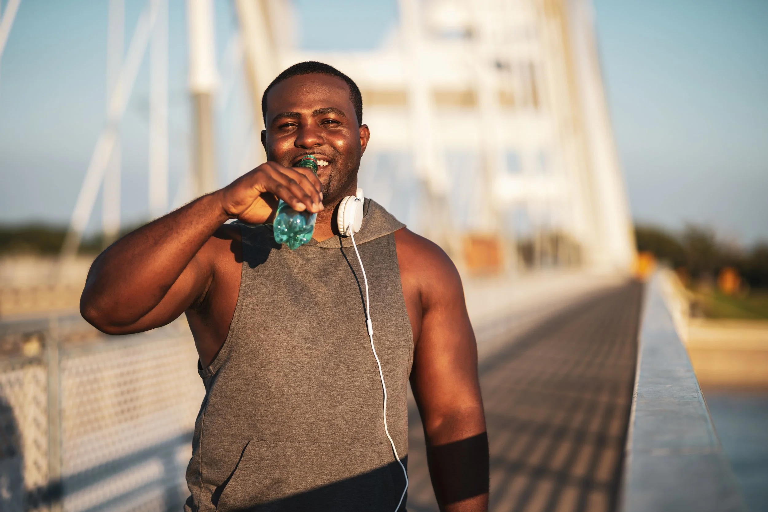 A man in athletic clothing holding a water bottle and smiling, with headphones around his neck, standing on a bridge during sunset.