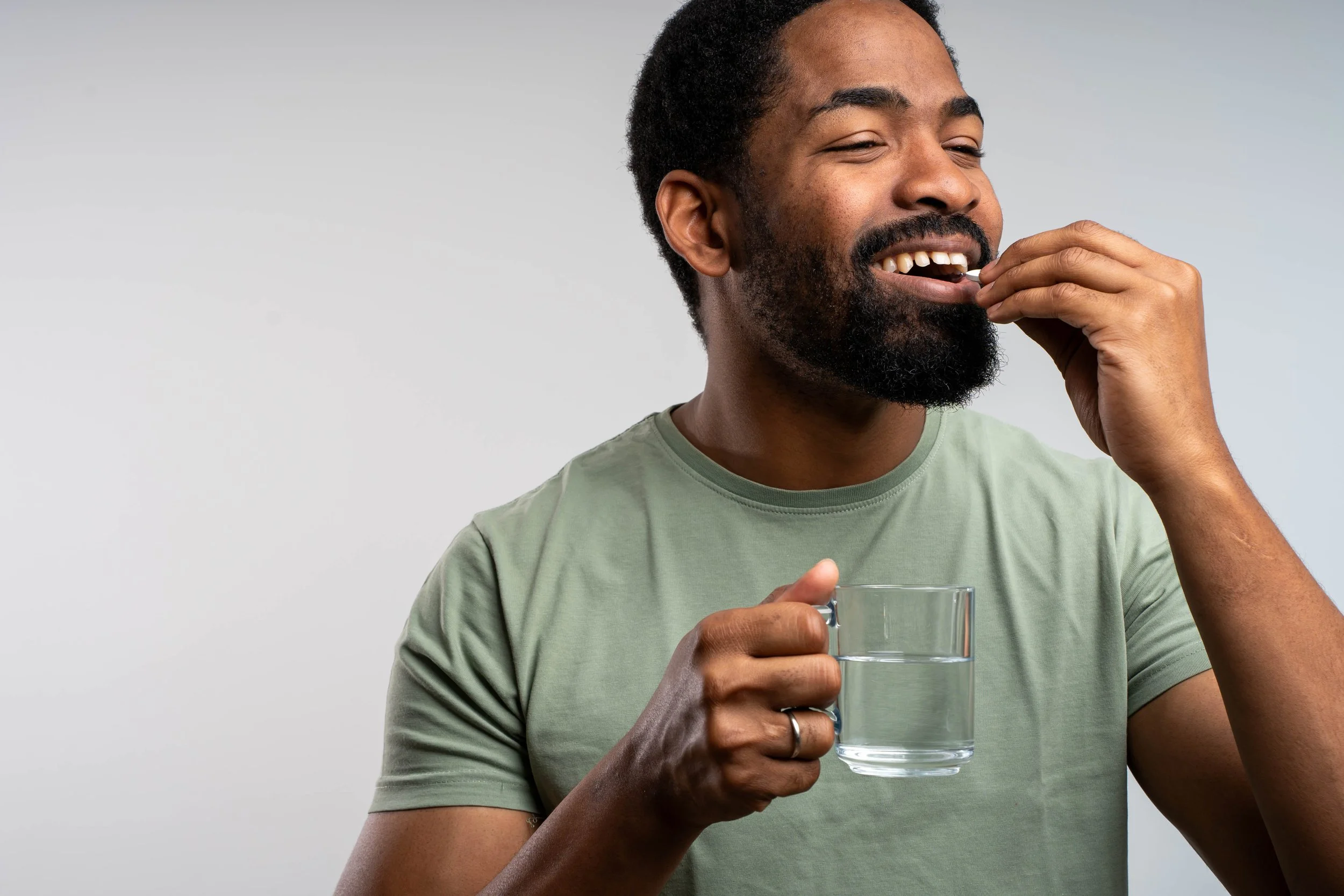 A man with a beard and short hair is smiling and holding a glass of water in his left hand while taking a pill with his right hand.