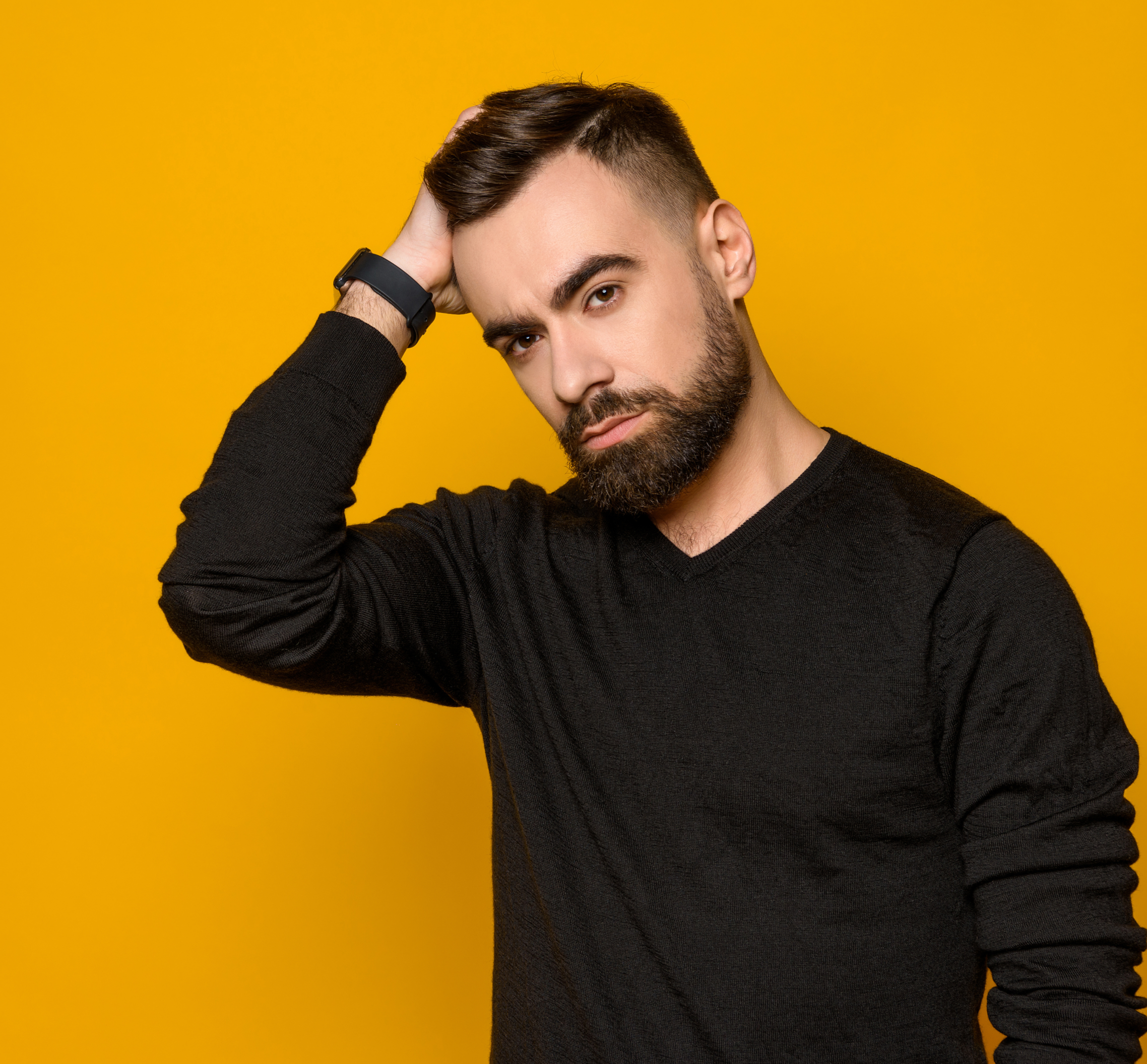 A man with a beard, wearing a black sweater and smartwatch, standing against a yellow background, looking at the camera with one hand on his head.