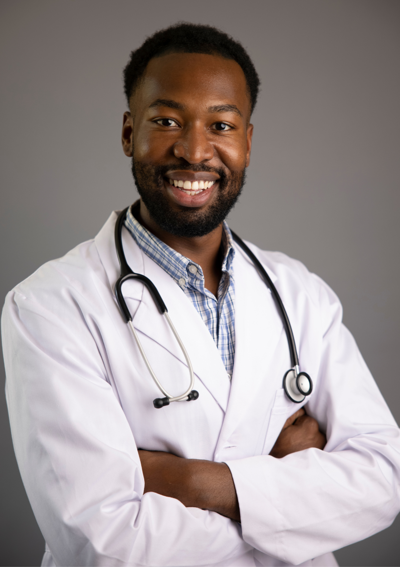 A smiling male doctor with a stethoscope around his neck, arms crossed, wearing a white coat and a checkered shirt against a gray background.