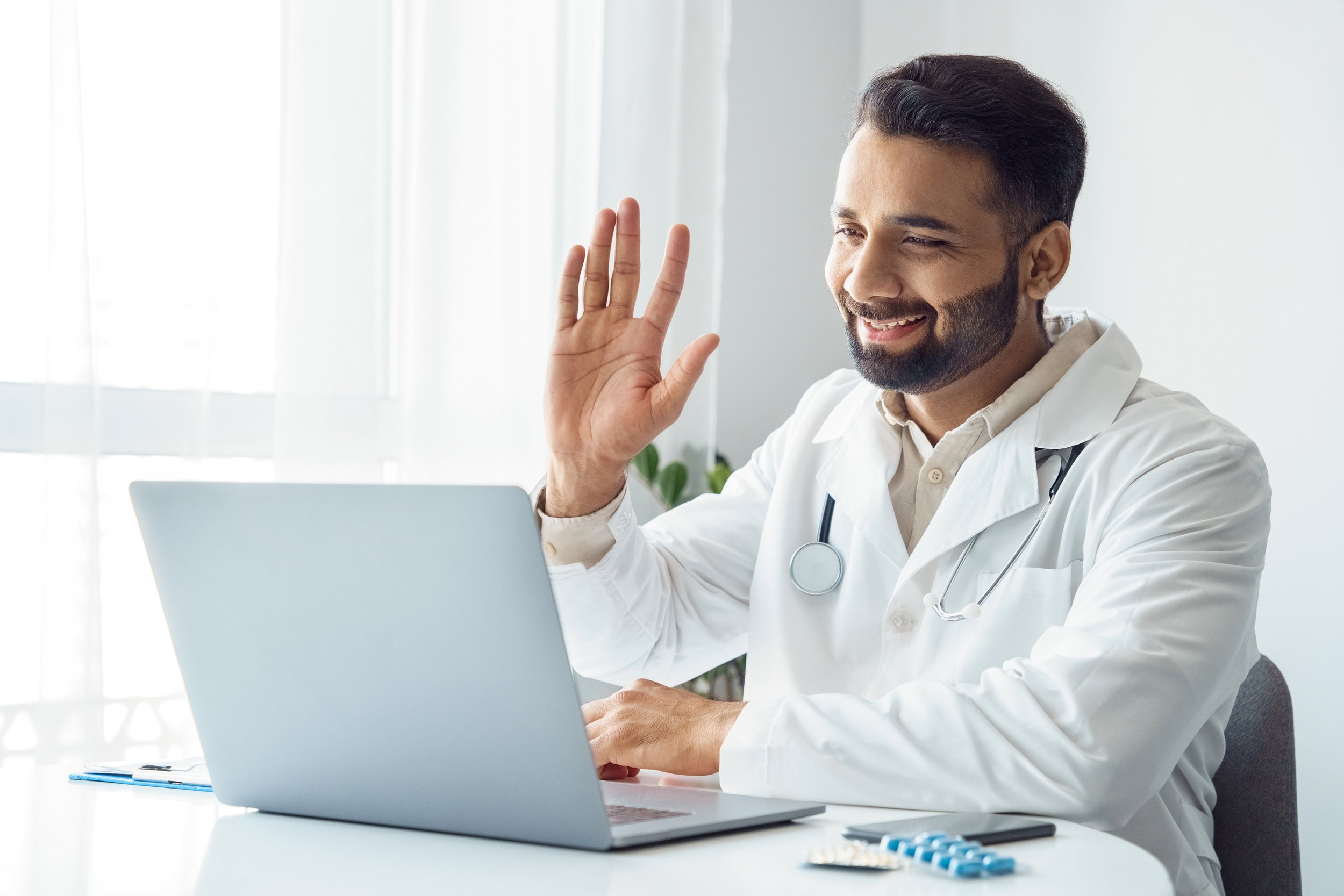 Male doctor in white coat and stethoscope smiling and waving while sitting at desk with laptop and medical supplies.
