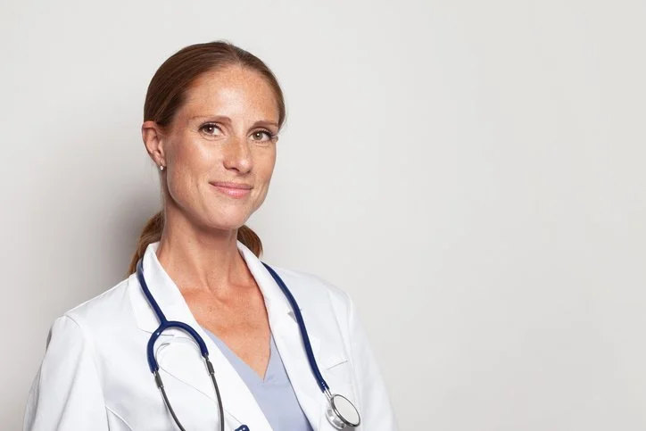 A female healthcare professional in a white coat with a stethoscope around her neck, standing against a plain light gray background.