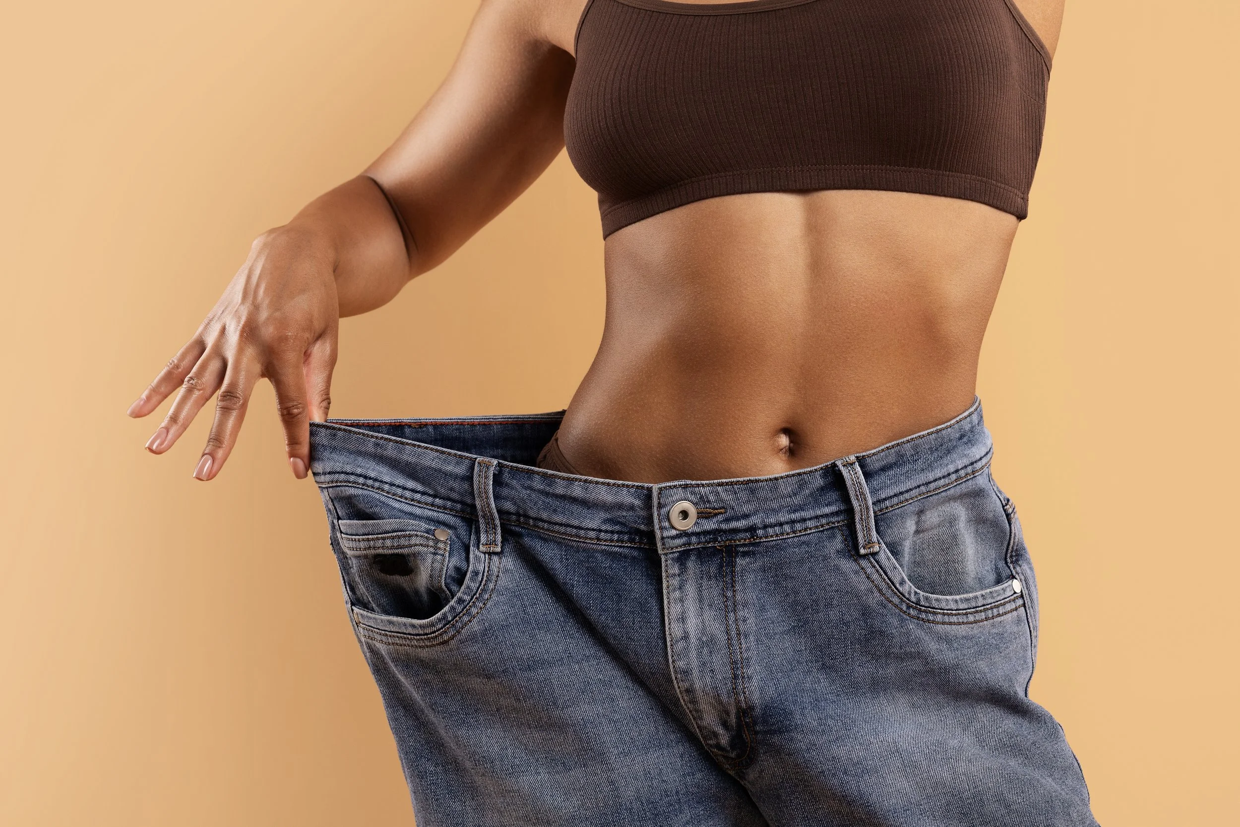 A woman wearing a brown crop top and oversized blue jeans with no pants, pulling the waistband out to show they are too big.