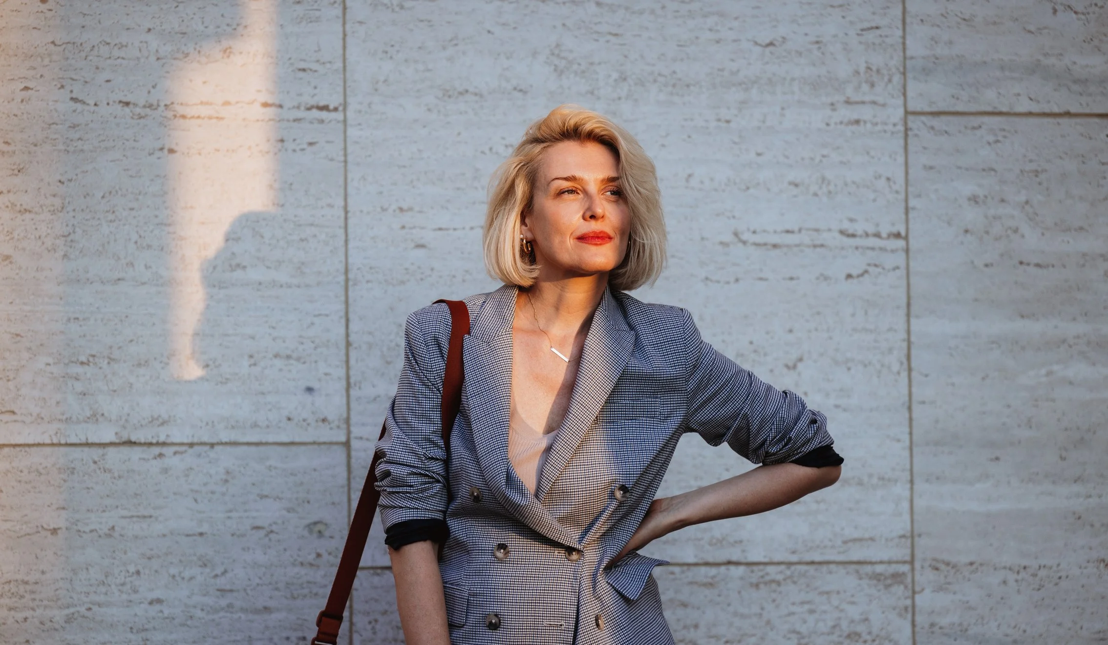 A woman with blonde hair wearing a plaid blazer, standing with one hand on her hip against a stone wall, looking thoughtfully into the distance.