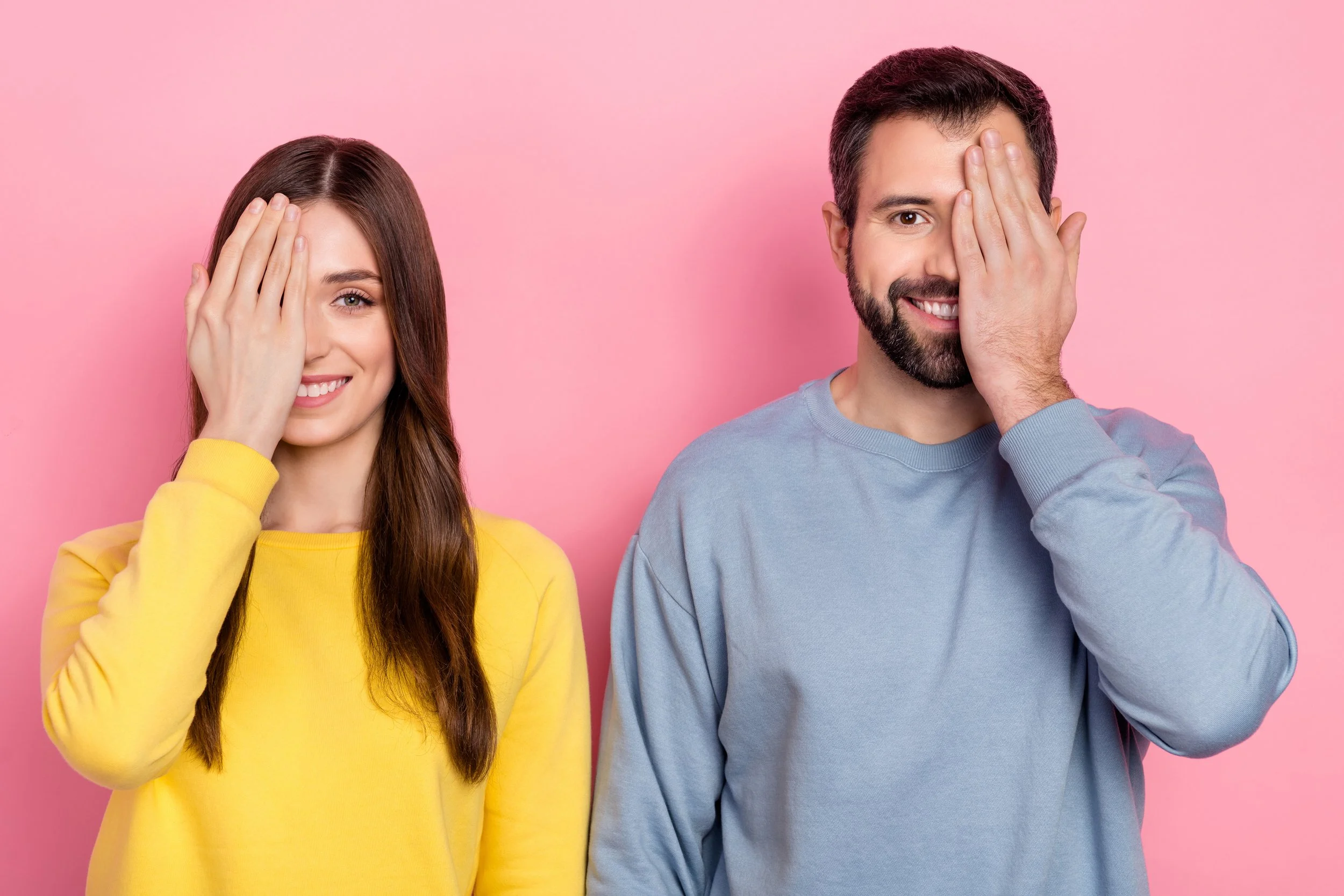 A woman in a yellow sweatshirt and a man in a light blue sweatshirt cover one eye with their hand, smiling against a pink background.