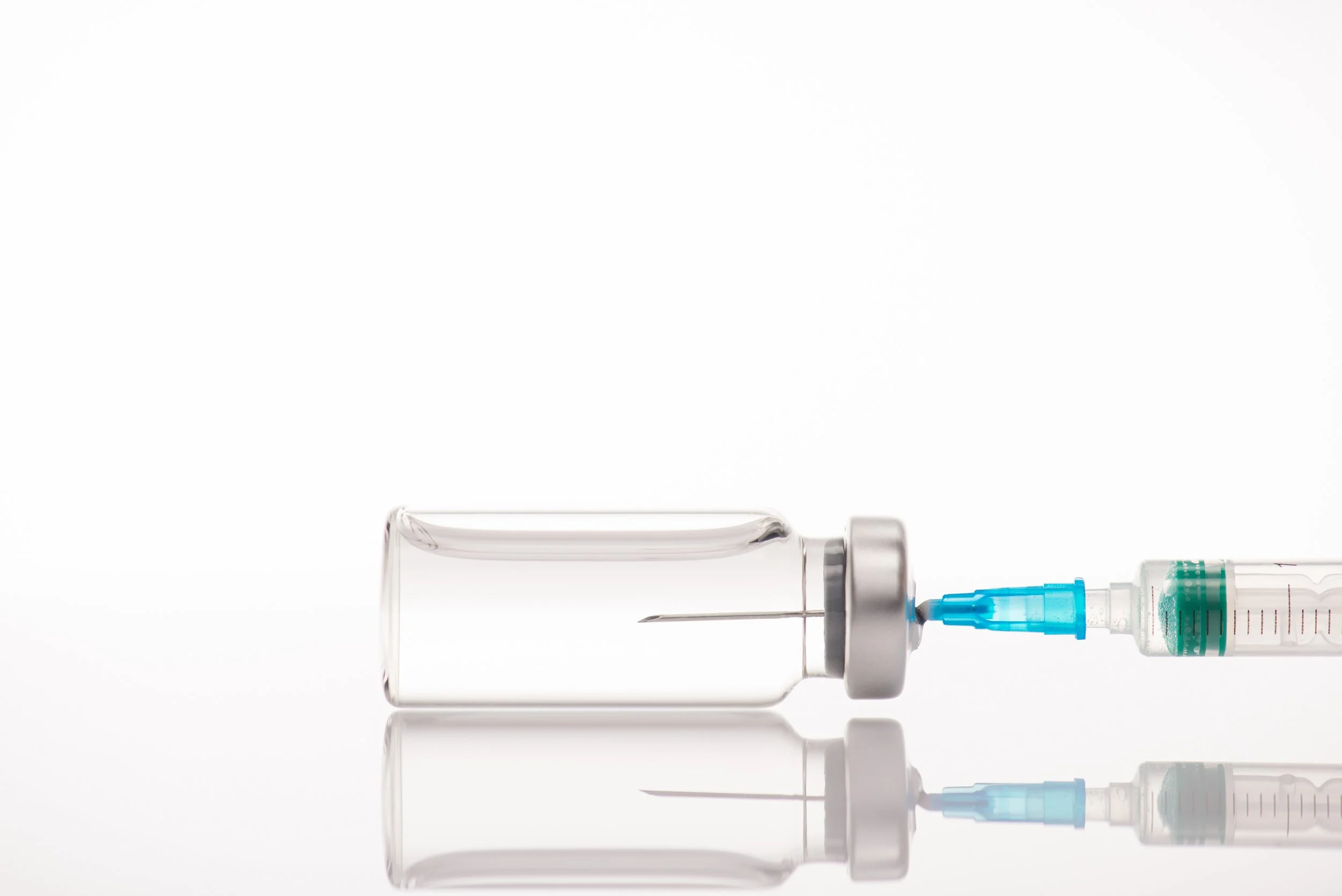 A medical syringe connected to a clear glass vial, lying horizontally on a reflective surface, with a white background.