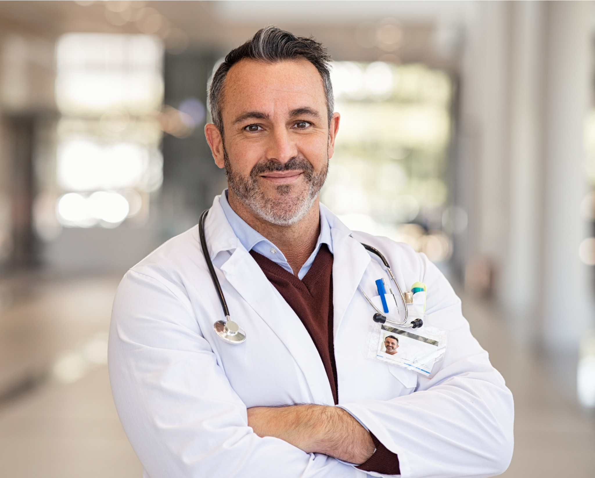 A male doctor with a stethoscope around his neck poses confidently in a hospital corridor with blurred background.